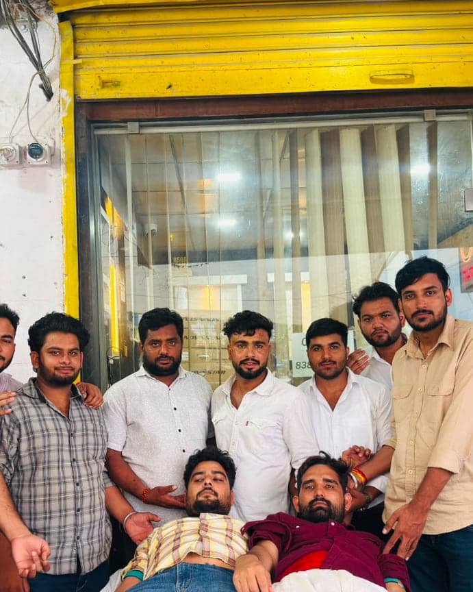 Two men donating blood side by side while a group of volunteers stands with them at the Allomart Pharmacy blood donation camp, Badpipli, Jaipur